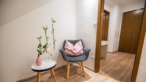 Una habitación con piso de madera, una pequeña mesa redonda con un jarrón rosa y una planta, una silla gris con cojín rosa, y una puerta de madera al fondo.