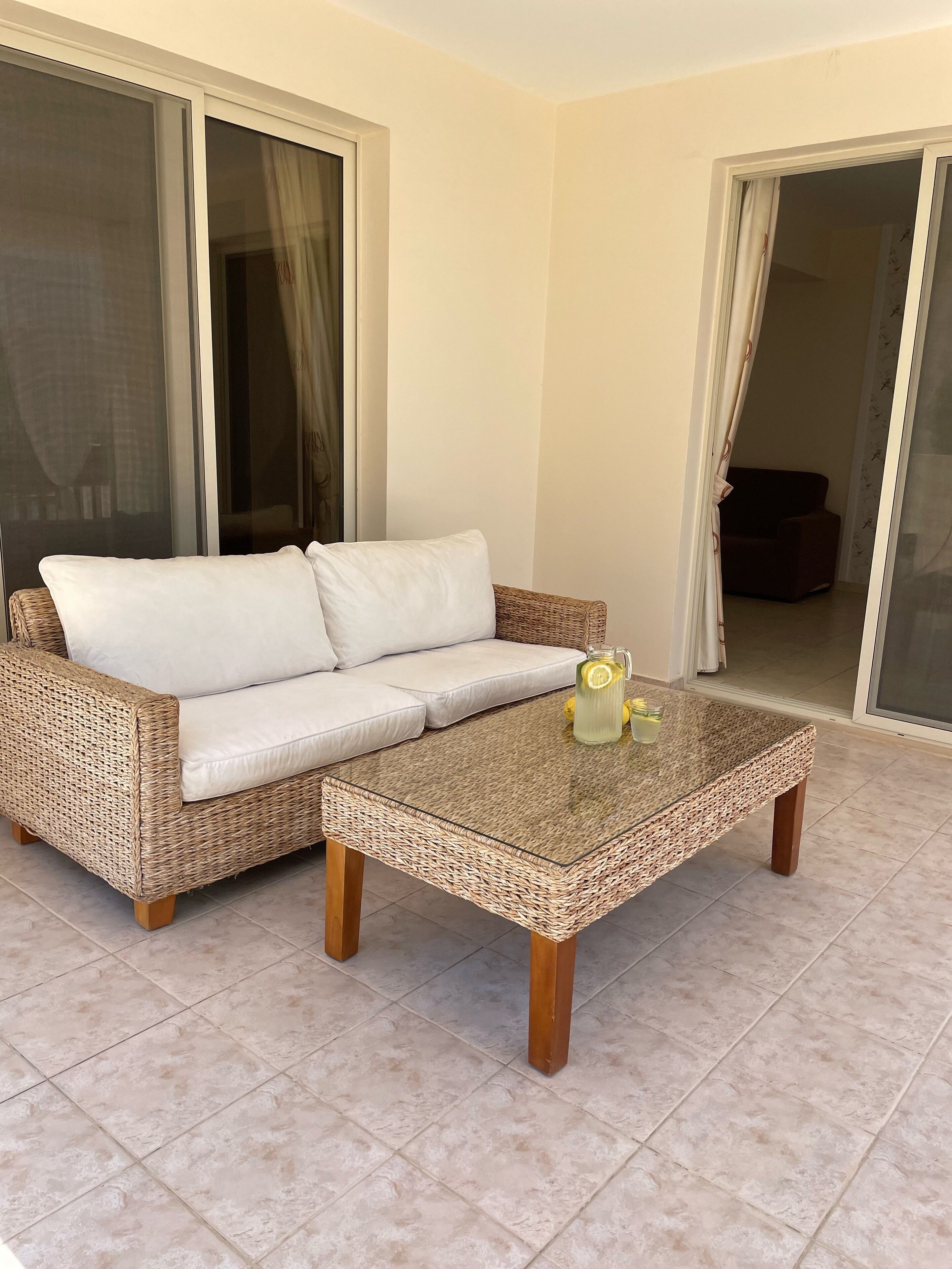 A wicker sofa with white cushions and a glass-top coffee table with a lemon pitcher on it.