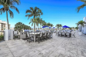 An outdoor patio with a grill, seating, and palm trees. - Regatta - William Raveis Rentals (Naples)
