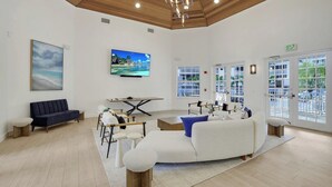 A spacious living area with a large TV, a sofa, a dining table, and a view of the outdoors through large windows. - Regatta - William Raveis Rentals (Naples)