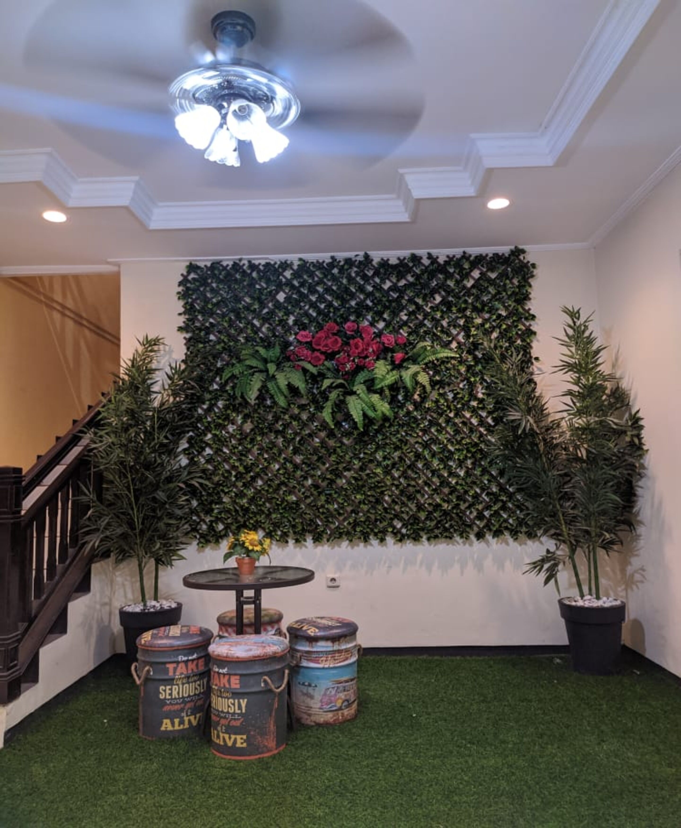 Una habitación con piso de césped artificial verde, una pared con plantas de flores rojas y un ventilador de techo. Hay plantas en macetas y latas de hojalata antiguas sobre el piso.