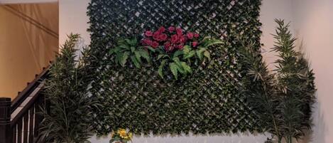 Una habitación con piso de césped artificial verde, una pared con plantas de flores rojas y un ventilador de techo. Hay plantas en macetas y latas de hojalata antiguas sobre el piso.