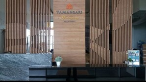 A reception area with a wooden panel wall displaying "TAMANSARI Mahogany APARTMENT" signage. There is a reception desk with a marble top and a potted plant. - Mahogany x Sagita Karawang by Zuzu (Karawang)