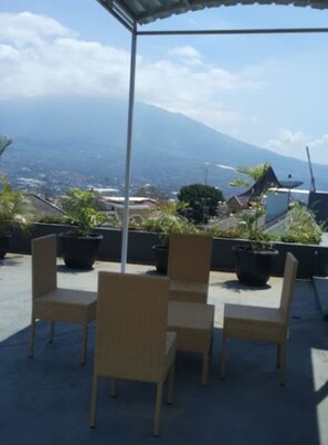 A covered outdoor seating area with wicker chairs and potted plants, overlooking a cityscape and a mountain. - Villa Asri (Batu)