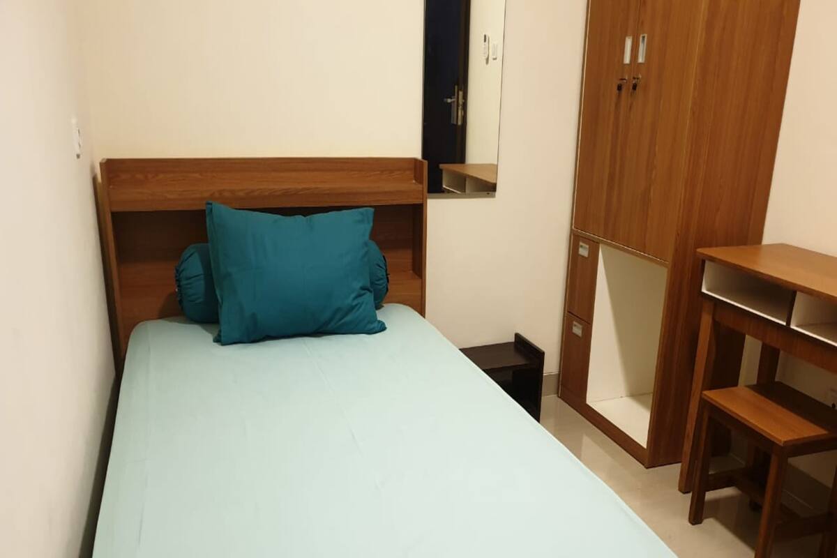 A single bed with a wooden headboard, a teal pillow, and a light blue bedspread in a room with a wooden wardrobe and a small desk.