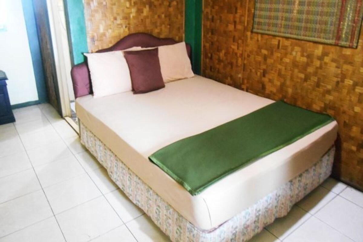 Ein Bett mit grüner Bettdecke und blumigem Bettzeug in einem Zimmer mit gewebter Wand und Fliesenboden.