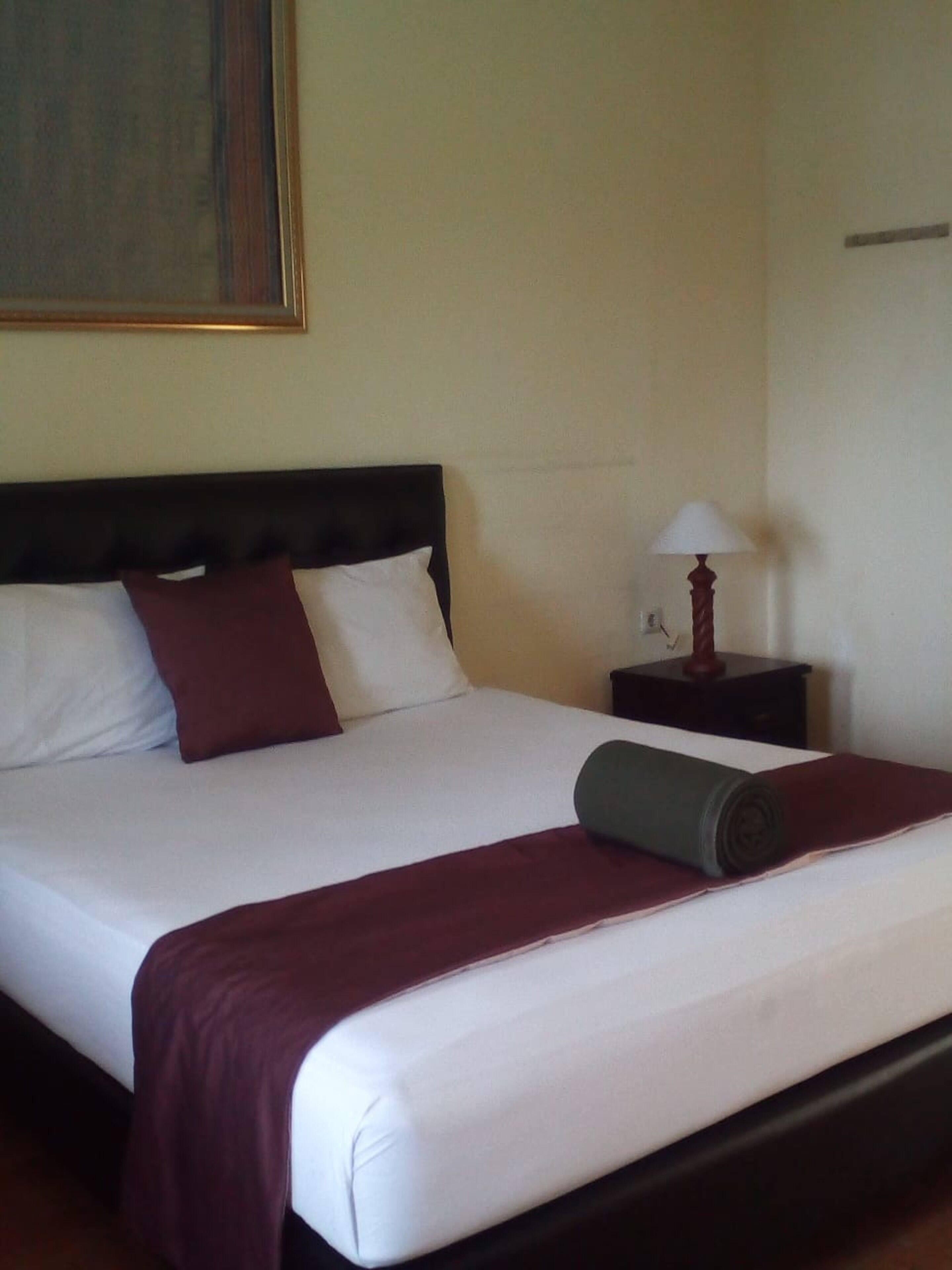 A neatly made bed with white and maroon bedding, a dark headboard, and a small bedside table with a lamp.