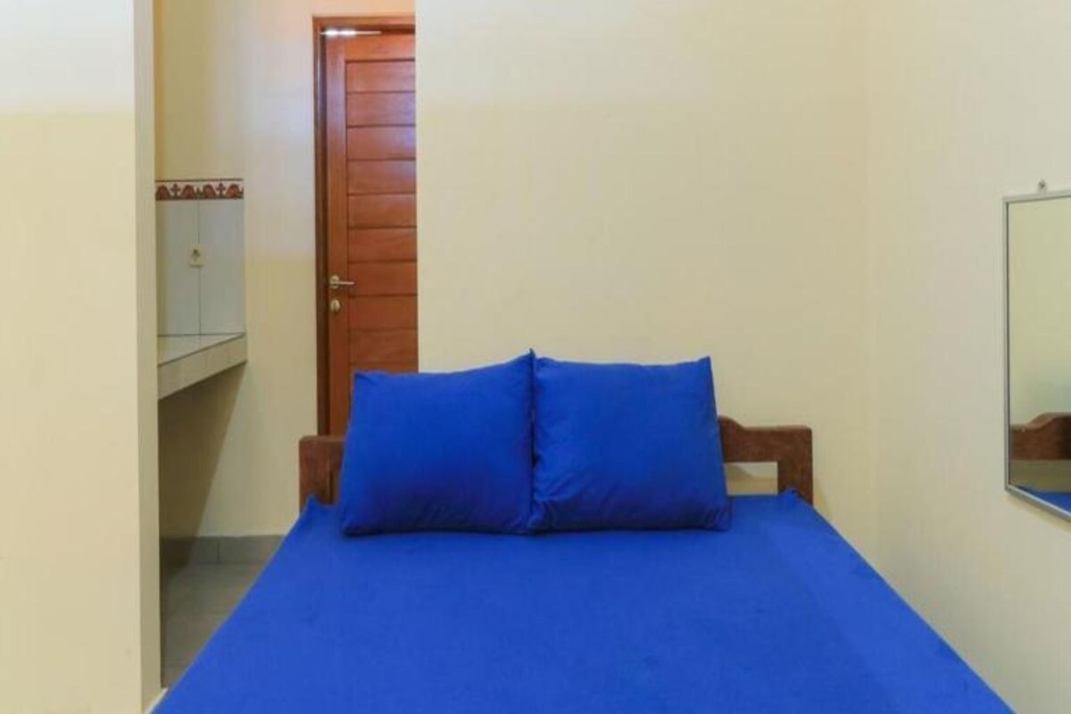 A room with a bed, a wooden door, a small cabinet, and a mirror on the wall.