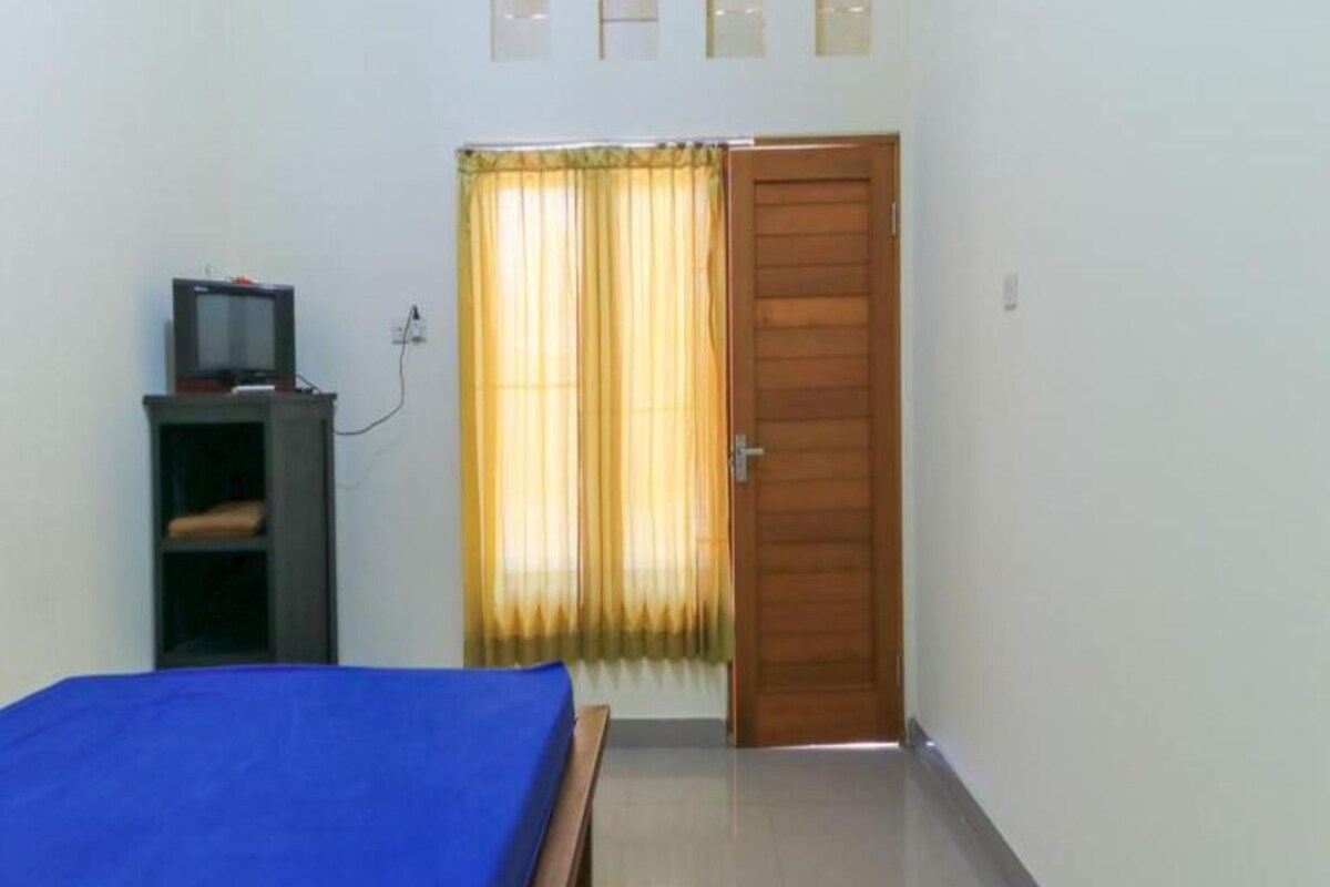 A room with a bed, a wooden door, a shelf with a television, and a window with curtains.