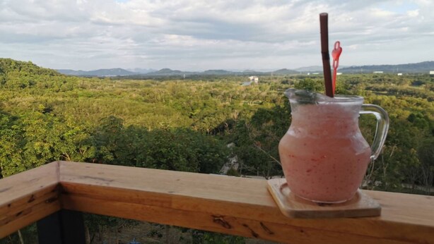 A wooden deck with a glass pitcher of pink drink on a coaster, overlooking a lush green landscape with distant mountains. - PP Residence Phuket (Sa Khu)