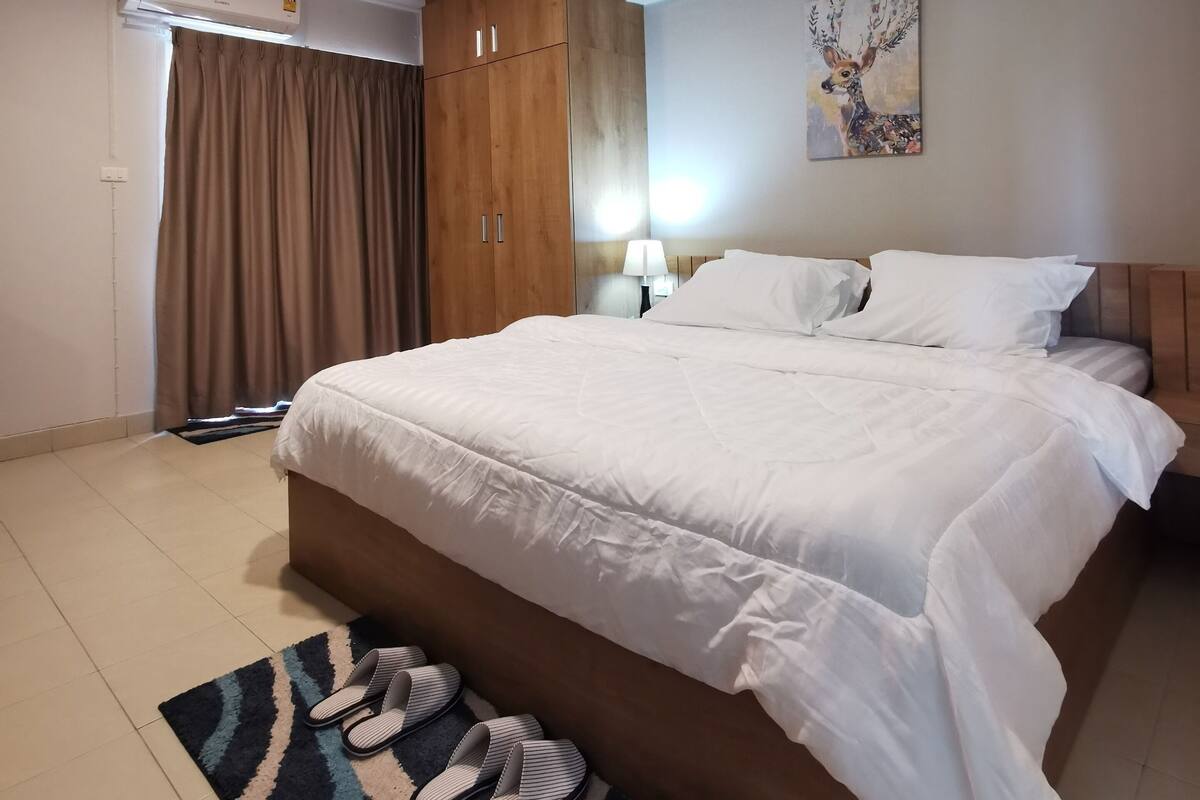 A hotel room with a large bed, a wooden wardrobe, an air conditioning unit, a painting on the wall, and a patterned rug.