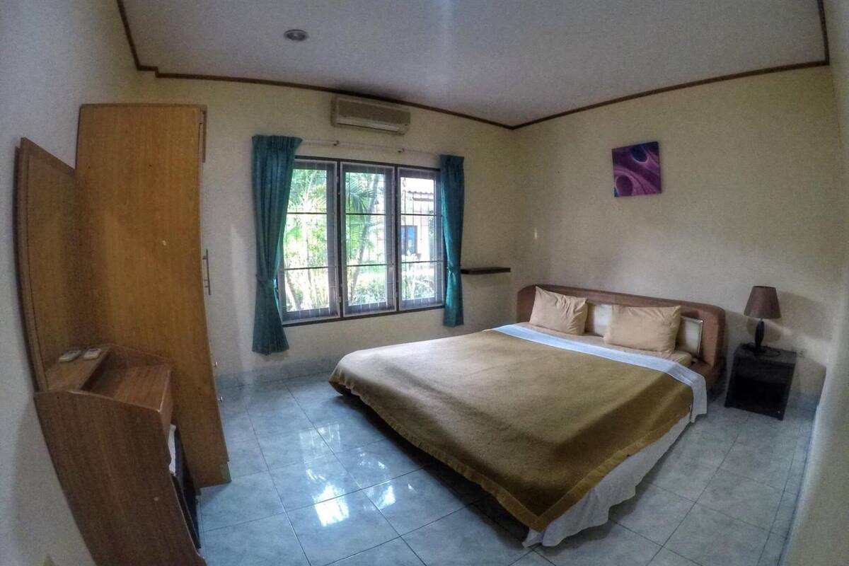 A bedroom with a bed, a nightstand, a desk, a window with curtains, and an air conditioning unit.