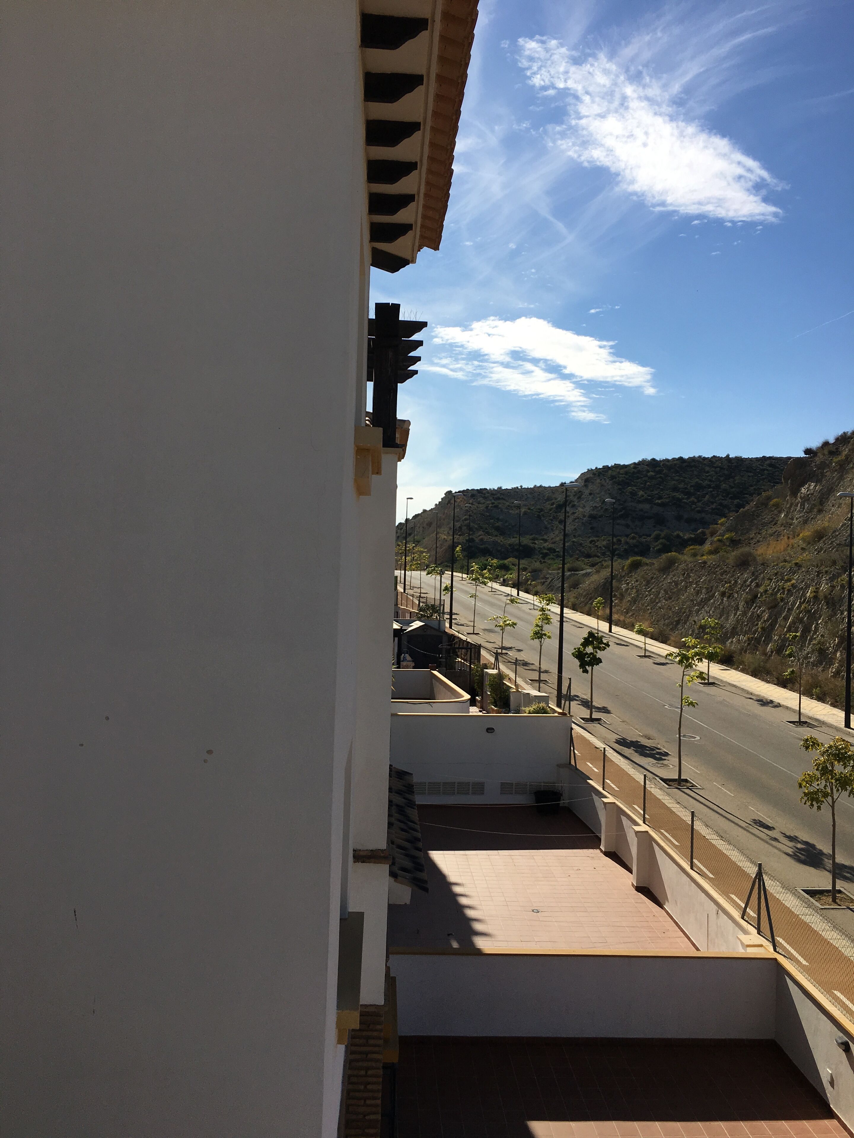 Vista des d'un balcó sobre un carrer amb arbres i un paisatge amb muntanyes a la distància.