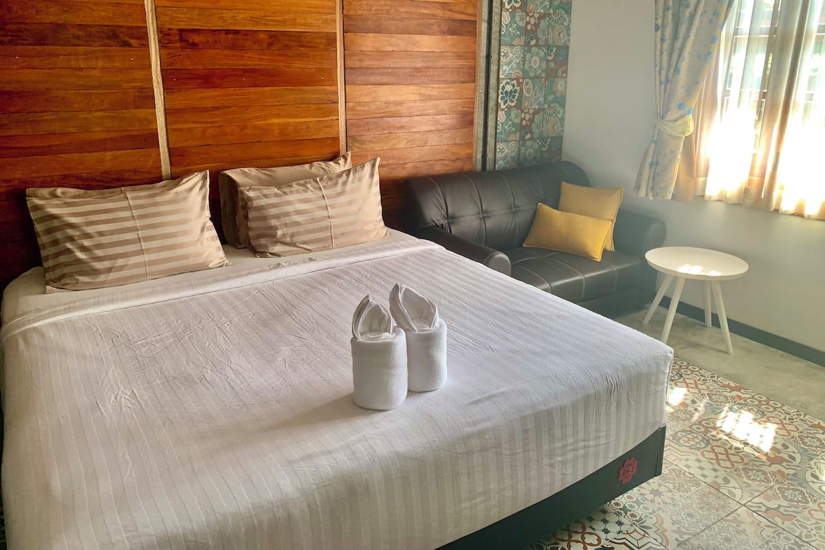 A neatly made bed with striped pillows, a leather sofa with a yellow pillow, and a small white table.