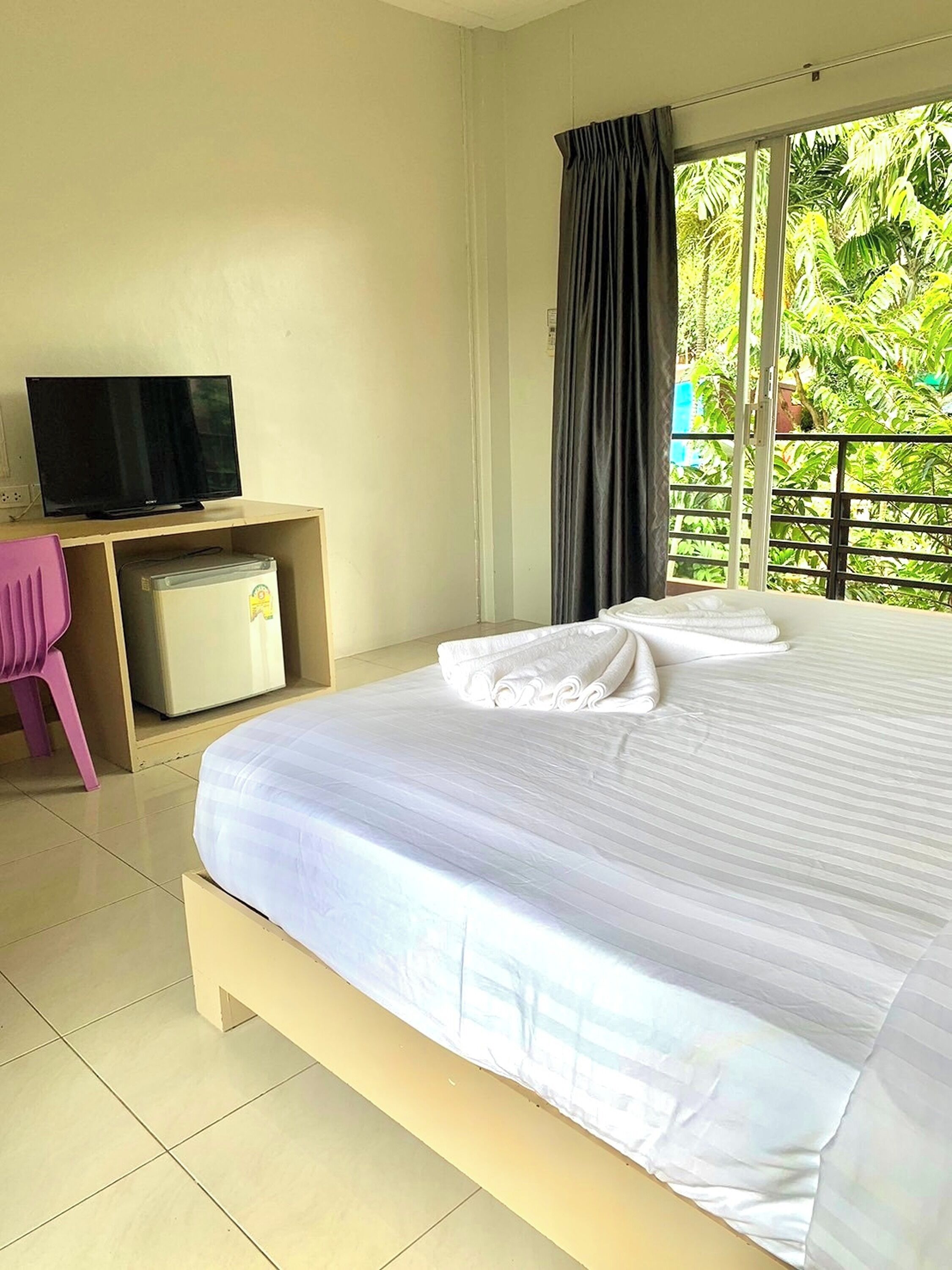 A hotel room with a bed, a television on a desk, a small refrigerator, a pink chair, and a balcony with greenery.