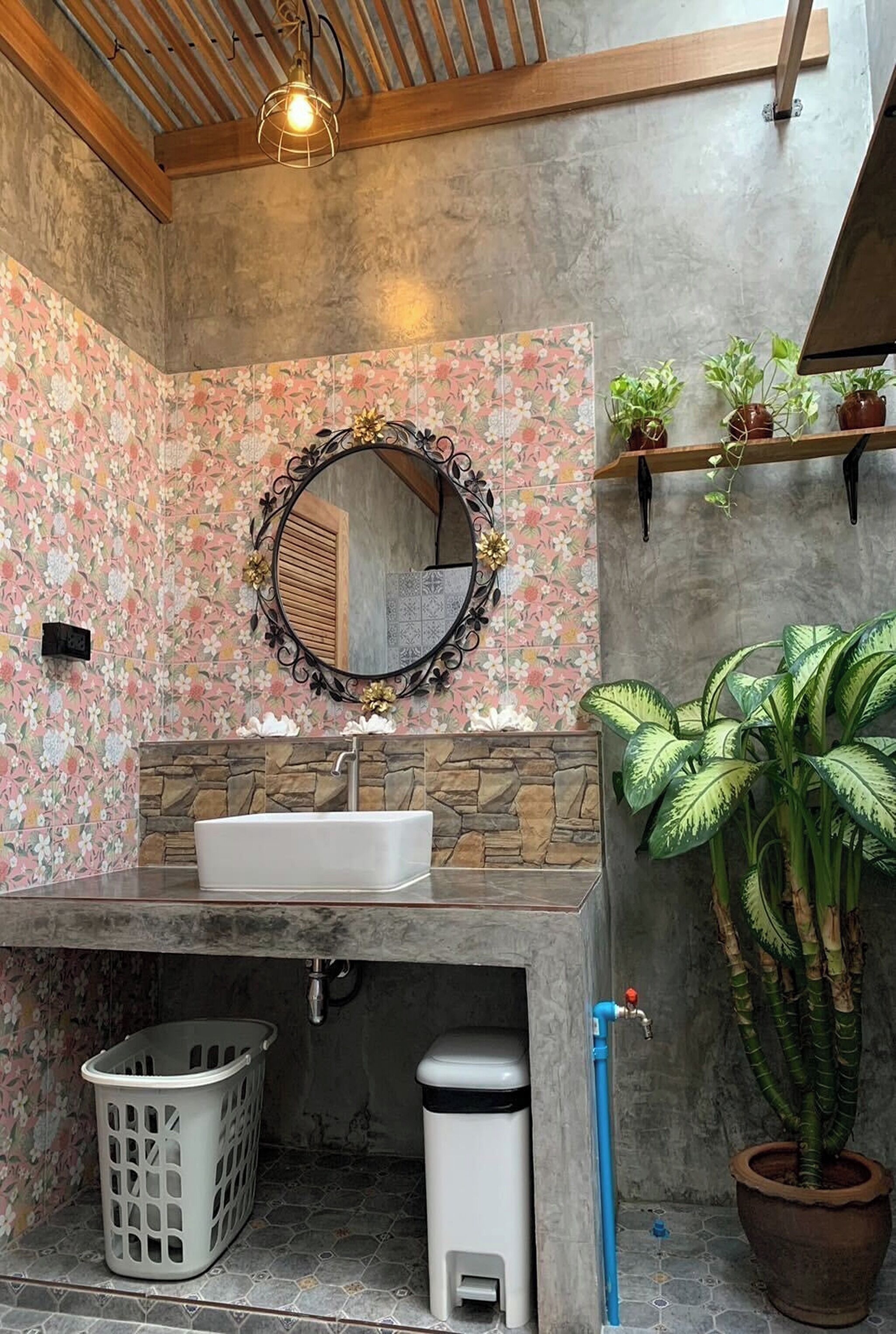 A bathroom with a stone sink, a round mirror, a laundry basket, a trash bin, and a potted plant.