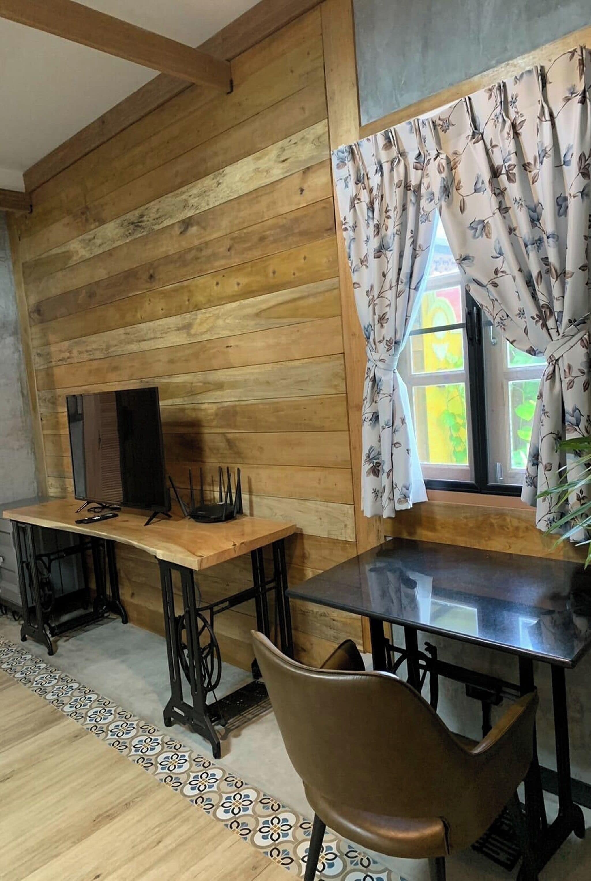 Une pièce aux murs en bois, avec un bureau en bois, une chaise, un téléviseur et une fenêtre ornée de rideaux à motifs floraux.