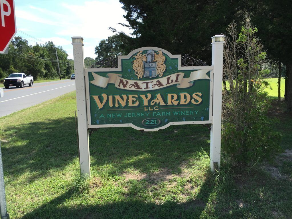A sign for Natali Vineyards LLC, a New Jersey farm winery, with a road sign and a stop sign in the background.