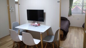 Una habitación pequeña y ordenada con una mesa blanca, cuatro sillas, un televisor de pantalla plana, un reloj y una ventana con cortinas.