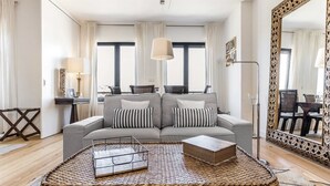 Ein modernes Wohnzimmer mit einem grauen Sofa, gestreiften Kissen, einem gewebten Couchtisch und einem großen, verzierten Spiegel.