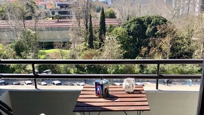 Ein Balkon mit zwei Holzstühlen und einem kleinen Tisch, mit Blick auf eine Stadtlandschaft mit Gebäuden und Bäumen.