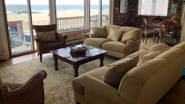 Een woonkamer met een groot raam dat uitkijkt over een strand, ingericht met beige banken en rieten stoelen.