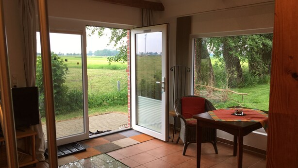 A room with a glass table, a wicker chair, and a view of a green field through large sliding glass doors. - Breekate vacation apartment Gustav - surrounded by greenery (Butjadingen-Tossens)