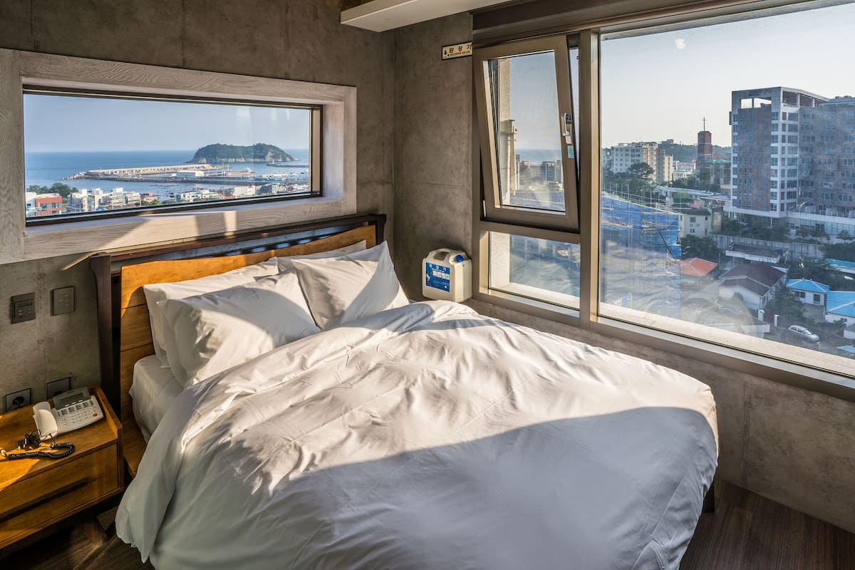 A hotel room with a large bed, a wooden headboard, a bedside telephone, and a window offering a city view.