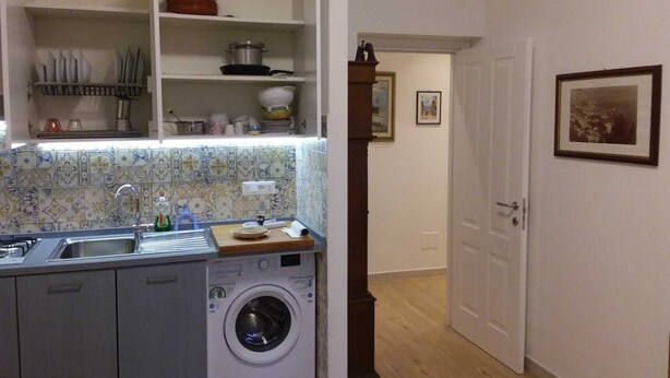 Une petite cuisine équipée d’un lave-linge, d’un évier et d’un petit plan de travail. Une porte donne accès à une autre pièce et un tableau encadré est accroché au mur.