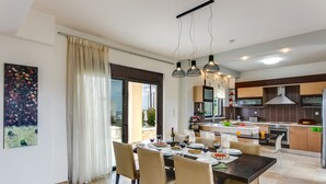 A modern kitchen with a central island, dining area with a table set for six, and a large window with curtains. - Villa Selena - Lux 4BD Residence (Chania)