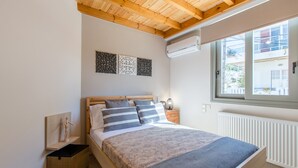 A bedroom with a wooden ceiling, a bed with a patterned bedspread, a nightstand with a lamp, and a window with a view of buildings. - Kismet Apt - Retreat to a Stylish home (Chania)