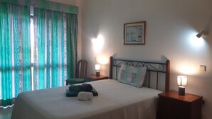 A neatly made bed with a quilt, two bedside lamps, a chair, and a wall-mounted picture. - 211- Amazing garden level Apartment - fantastic location (Albufeira)