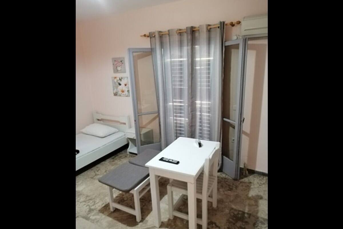 Ein kleines Zimmer mit einem Bett, einem Tisch mit Stuhl, einem Fenster mit Vorhängen und einer Tür.