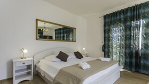 Ein ordentlich gemachtes Bett mit einer beigen Decke, zwei Nachttische mit Lampen, ein großer Spiegel und ein Fenster mit tropischen Vorhängen.