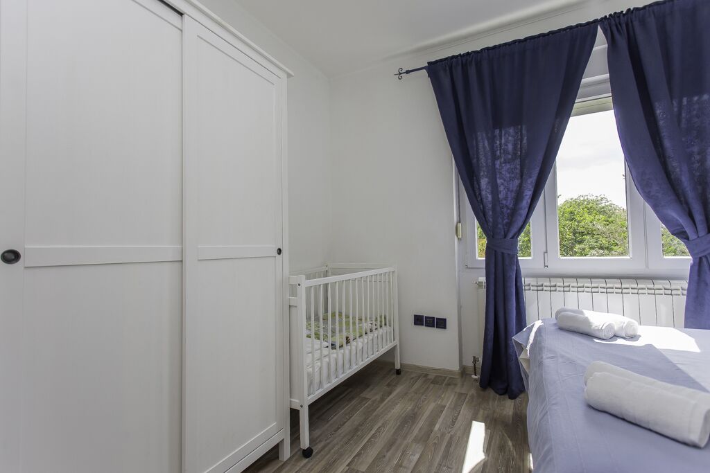 Un dormitorio con una cuna blanca, una cama con ropa de cama blanca y una ventana con cortinas azules.