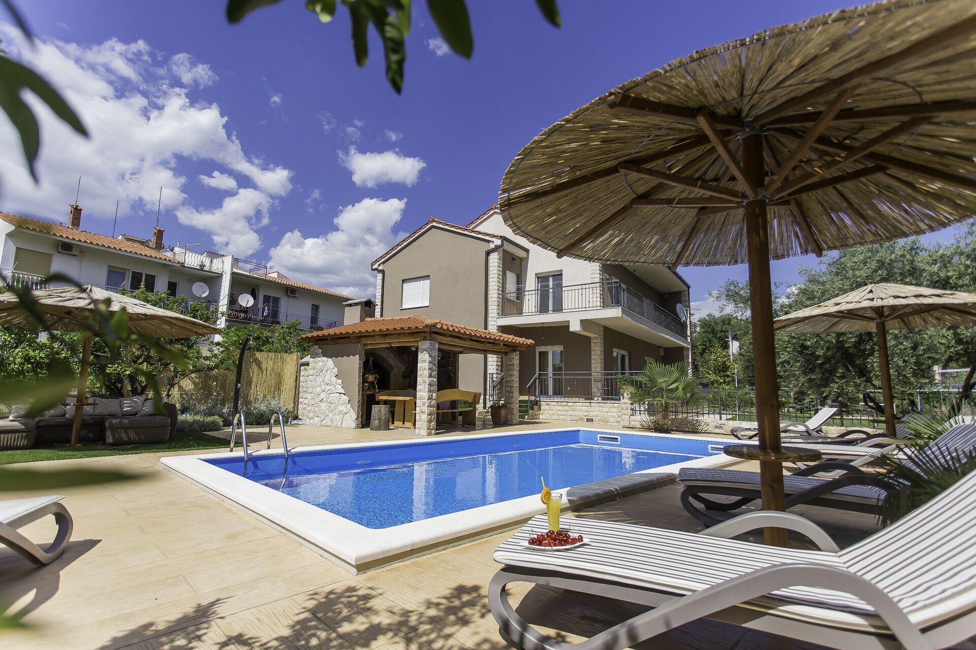 A swimming pool area with lounge chairs and umbrellas, surrounded by buildings with balconies and a stone wall.