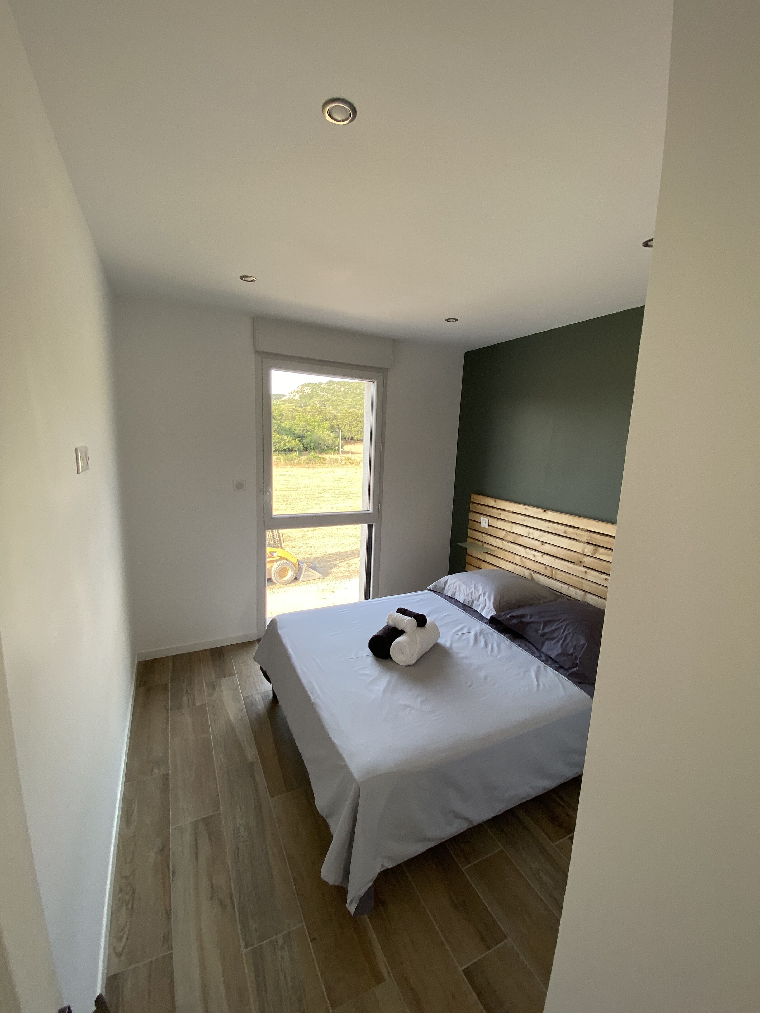 Une chambre à coucher avec un lit, une tête de lit en bois, une fenêtre et une vue sur la verdure à l’extérieur.