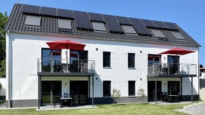 A modern two-story building with a flat roof, solar panels, and a balcony with red umbrellas.