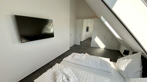 A modern hotel room with a large TV mounted on the wall, a bed with white bedding, and a skylight providing natural light.