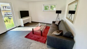 A modern living room with a grey sofa, a red rug, a glass coffee table, a TV on a white stand, and a large mirror on the wall.