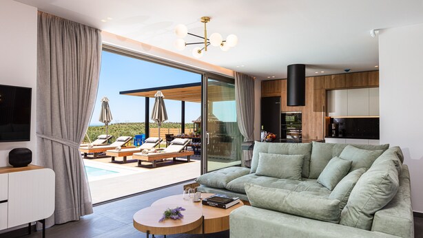 Ein modernes Wohnzimmer mit einer großen Couch, einem Couchtisch und einem Fernseher. Eine Glastür führt zu einem Poolbereich mit Liegestühlen und Sonnenschirmen.