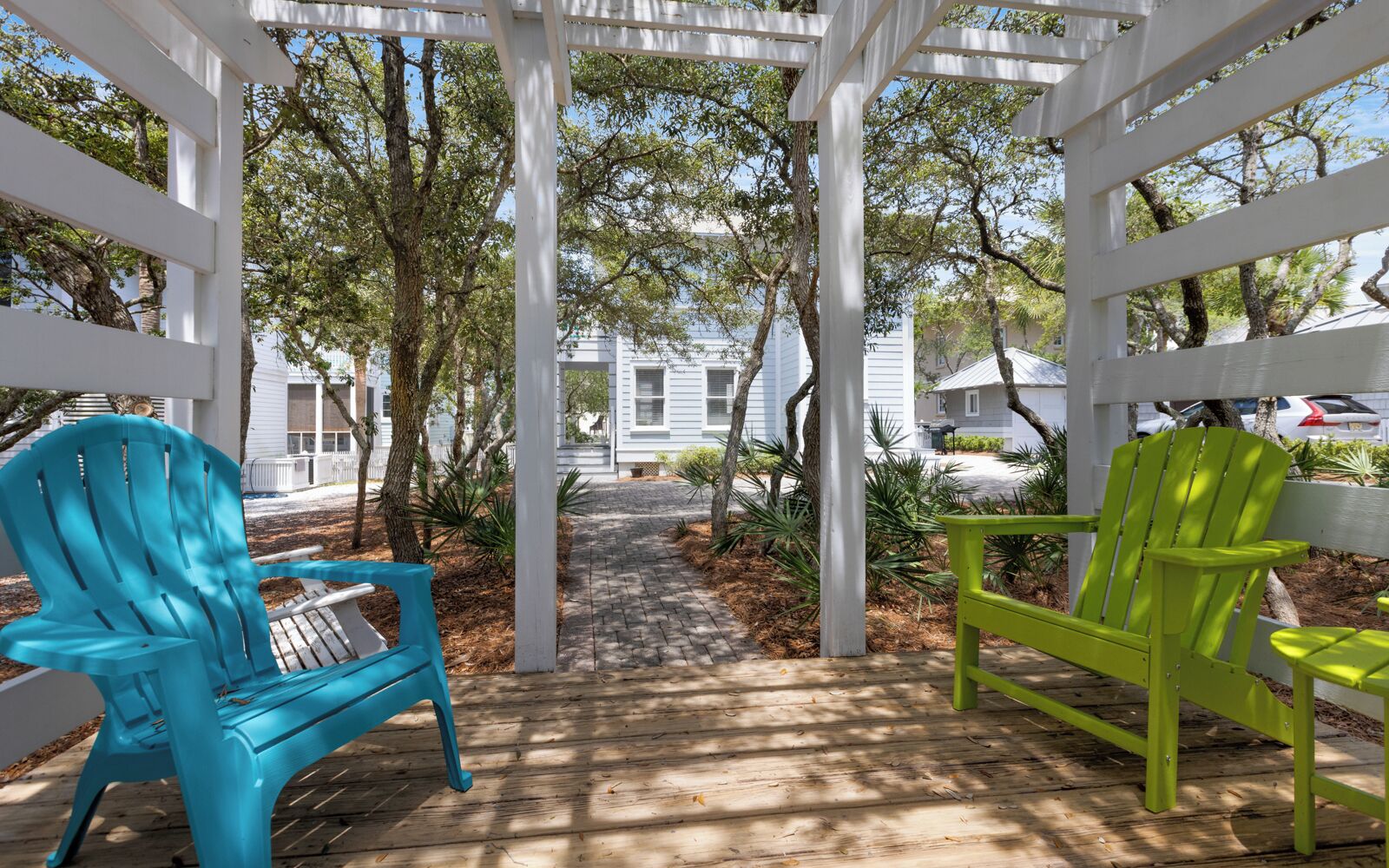 A covered outdoor area with a wooden deck, two Adirondack chairs, and a pathway leading to a building.