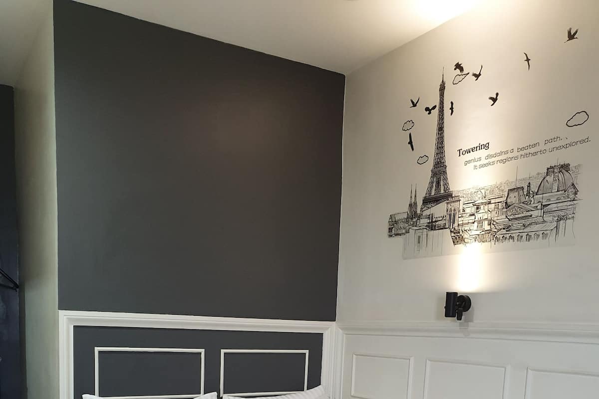 A neatly made bed with white bedding and a black pillow with "hello" text, a white headboard, and a wall with a cityscape illustration and text.