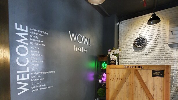 Zona de recepción con un letrero que dice 'BIENVENIDOS' y un letrero que dice '¡WOW! hotel' en varios idiomas. Hay un mostrador de recepción con un letrero que dice 'RECEPTION' y un reloj en la pared.