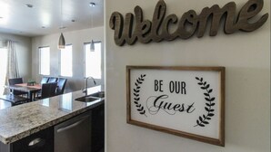 Une cuisine moderne avec un comptoir en granit, un coin repas avec une table et des chaises, et une pancarte « Bienvenue » au-dessus d’une affiche encadrée portant l’inscription « Soyez nos invités ».