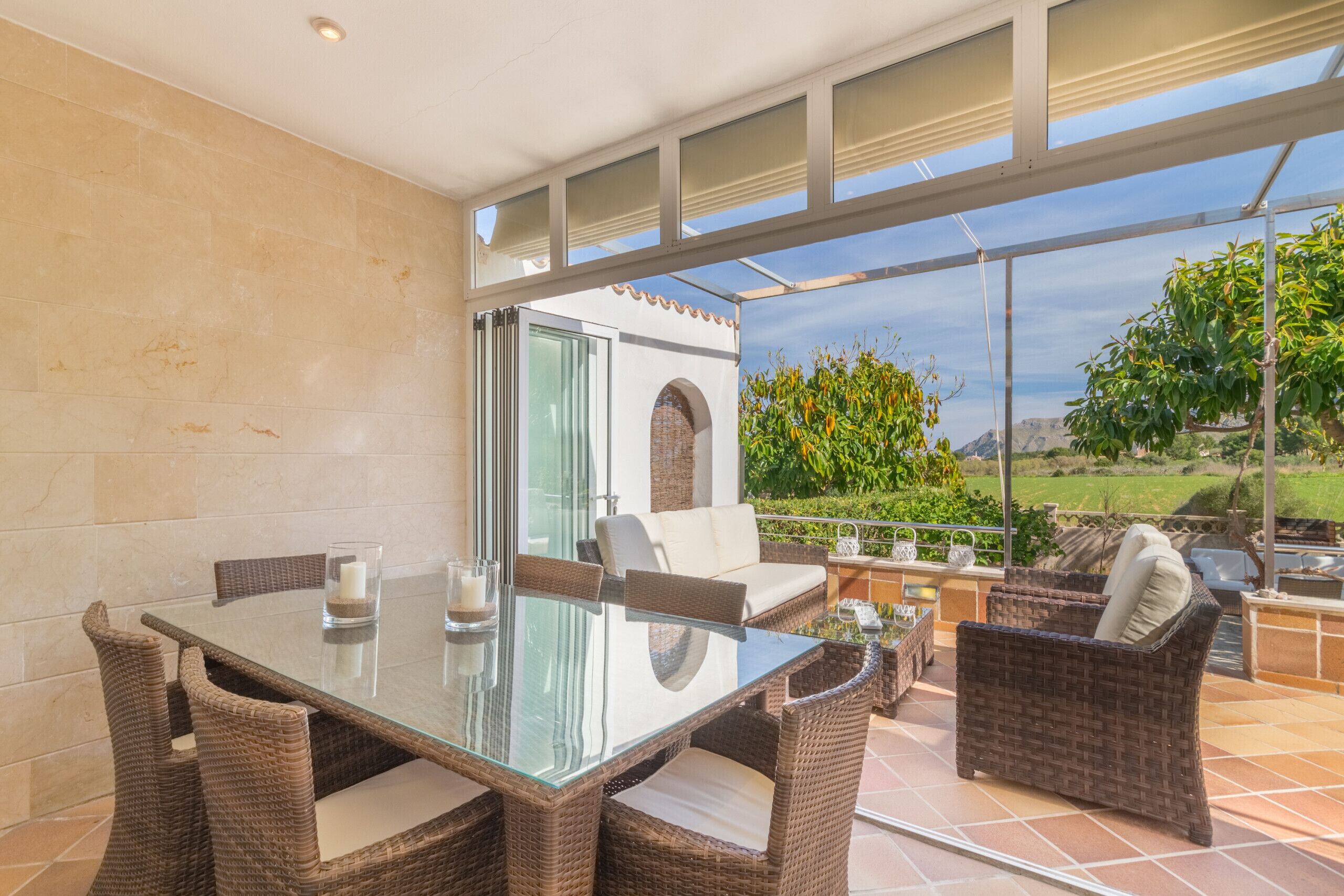 Mesa de jantar com tampo de vidro, cadeiras de vime, castiçal de vidro e vista para um jardim através de uma porta de vidro.