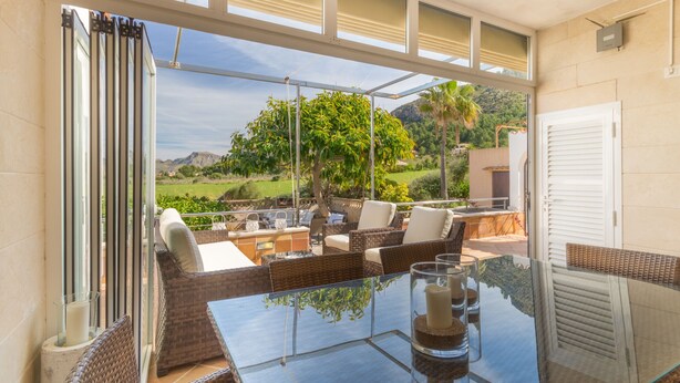 Un patio iluminado por el sol con mobiliario de mimbre, una mesa de vidrio y vistas a un paisaje verde.