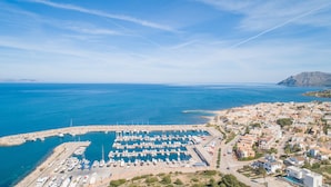 Un puerto deportivo con numerosas embarcaciones amarradas, un pueblo costero con edificios, y un cielo azul despejado.