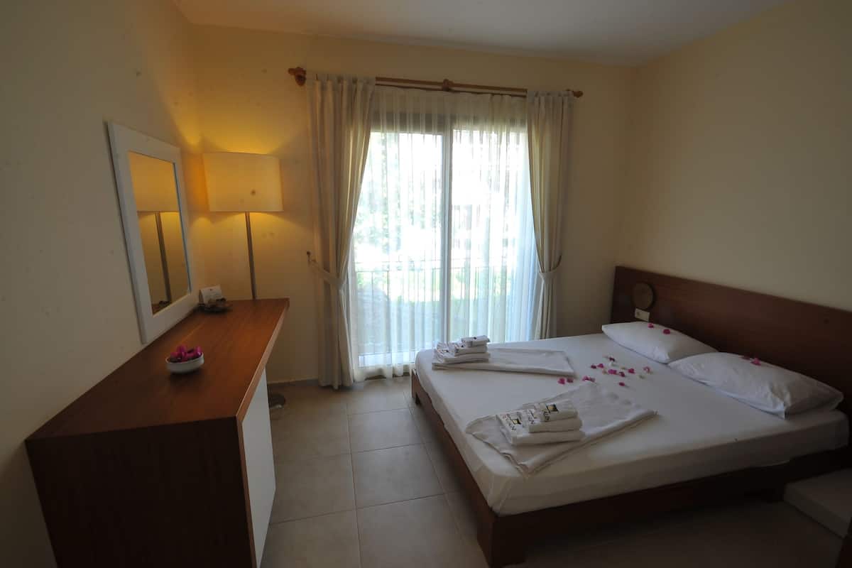 A hotel room with a bed, a wooden desk, a mirror, and a window with curtains.