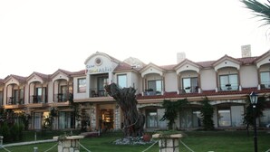 A hotel with a Mediterranean architectural style, featuring a central courtyard with a large tree and a fountain, surrounded by a chain-link fence.